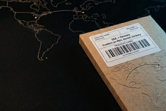 Flacher Karton aus Kraftpapier auf einer schwarzen Fläche mit eingeprägter Weltkarte, darauf ein Versandetikett mit der Aufschrift USA nach Germany, Frankfurt am Main, Hessen, Germany und einem Strichcode